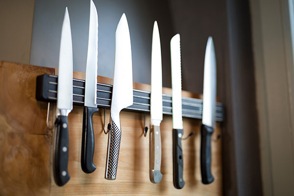A variety of 6 kitchen knives on a black knife magnet mounted on a wooden panel.