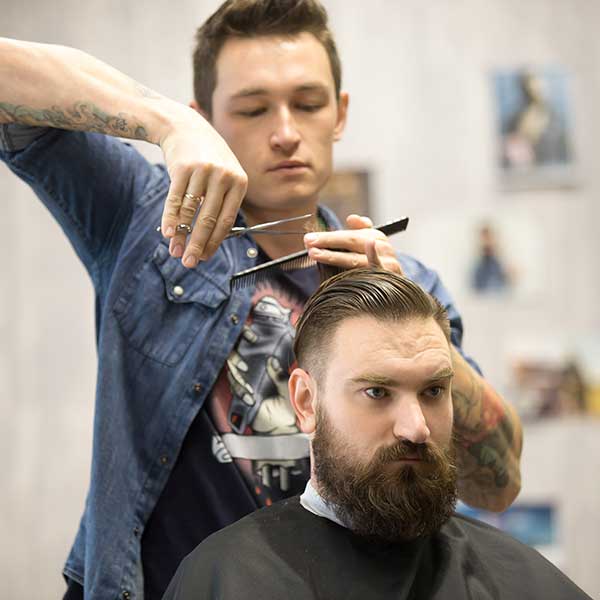 Front face view of a barber with shears and comb, cutting the hair of a bearded man. Man wears a black smock.