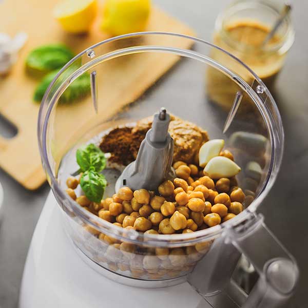 Top view. A food processor bowl with garbanzo beans, tahini, garlic, and basil, next to a cutting board with lemons and basil