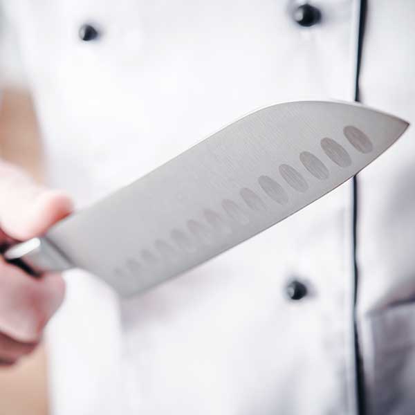 A close-up view, focused on a santoku knife, held by a chef wearing a white chef coat with black buttons