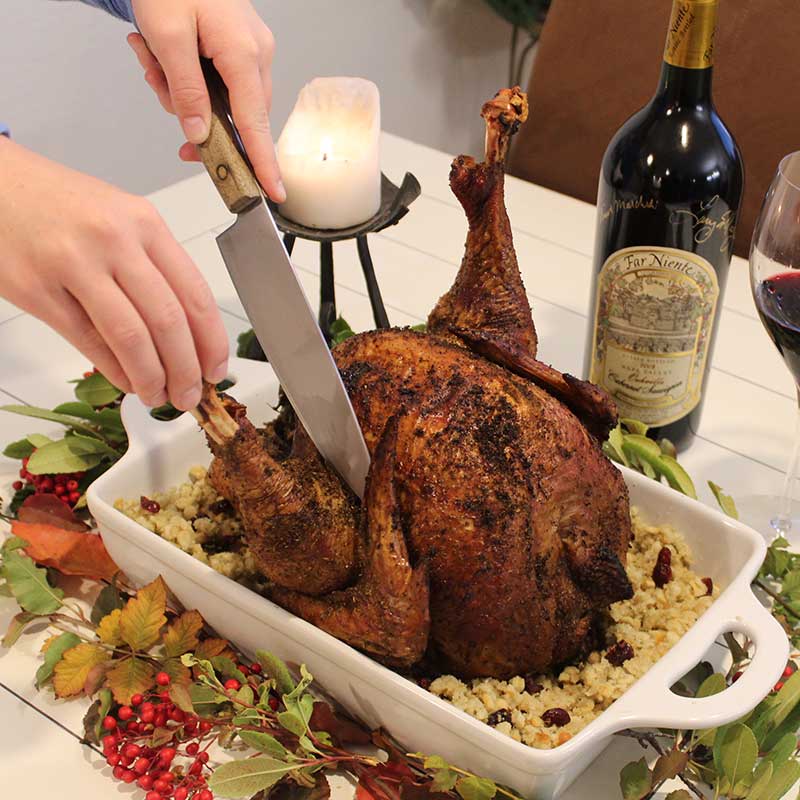 Mail-in-Knife-Sharpening-California Person begins to cut into thanksgiving turkey leg on a dinner table. Fall greenery, a candle, and wine decorate the table.