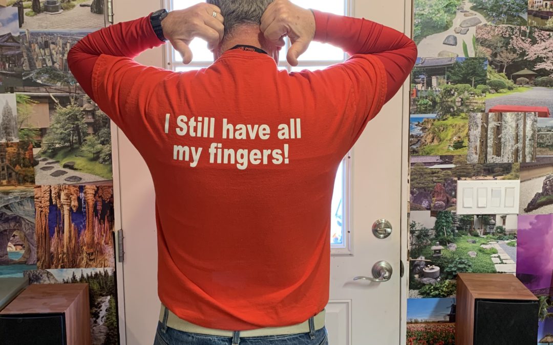 Man standing facing away with thumbs pointing down from his ears, wearing a red shirt saying "I still have all my fingers!"