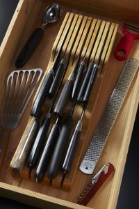 Knives in a bamboo knife drawer organizer in a drawer with some other cooking utensils.